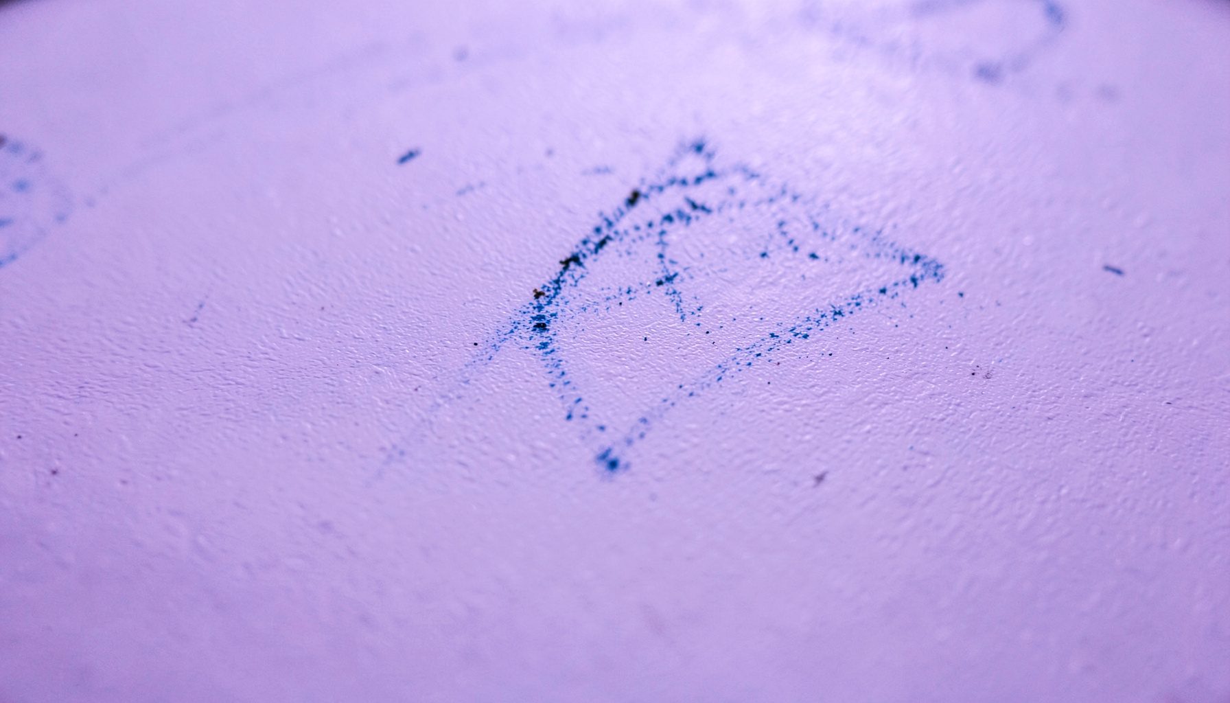 Extreme close-up photograph of a whiteboard surface with faint dry-erase marker smudges in blue and black, showing the textured matte finish of the board under soft overhead light.