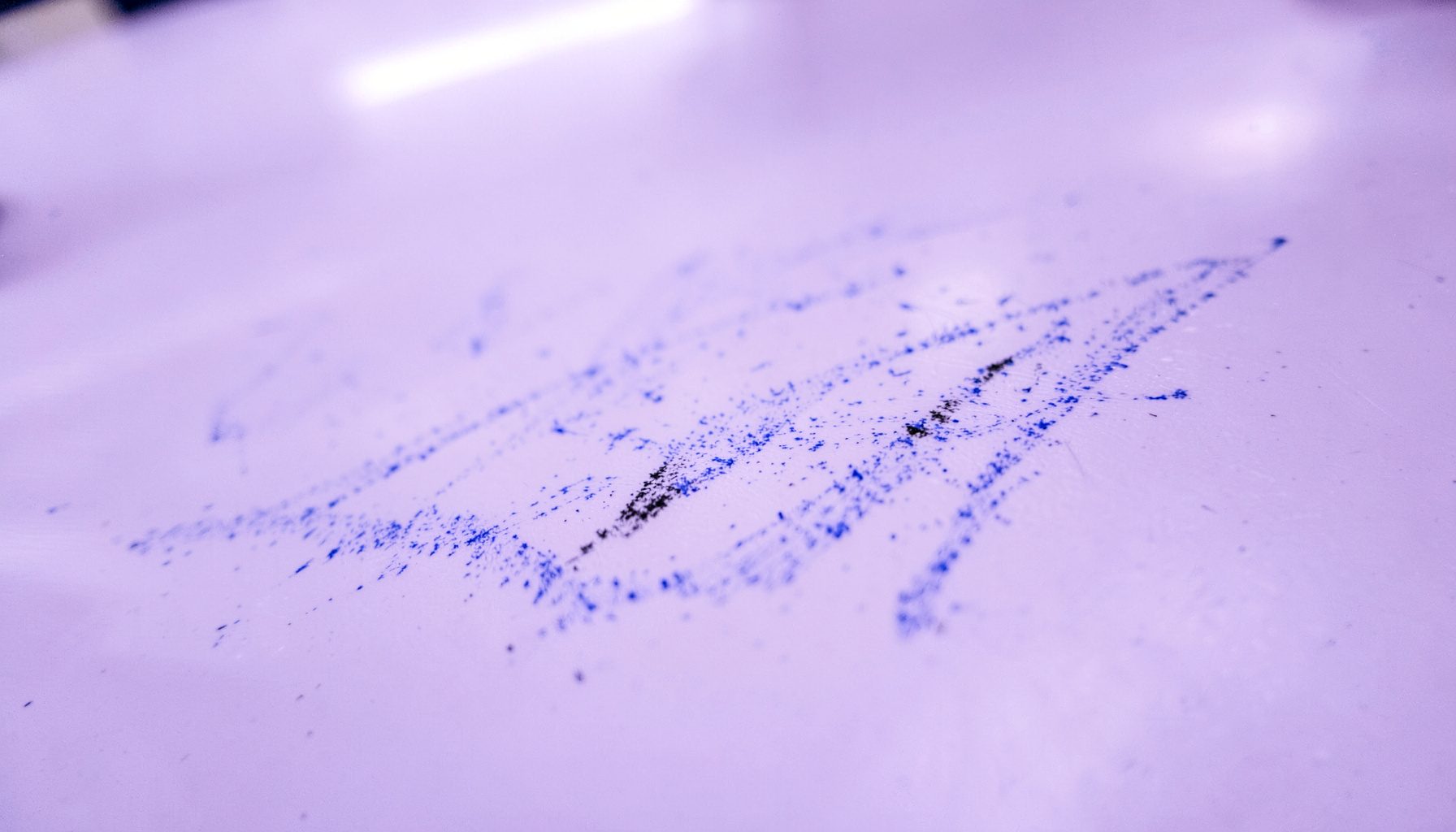 Extreme close-up of a whiteboard surface showing faint dry-erase marker smudges in blue and black against a glossy white background, with soft overhead fluorescent light creating a subtle gradient